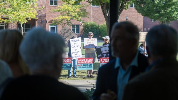 People speaking inside McCormick Center at the rededication with protestors in the background outside.